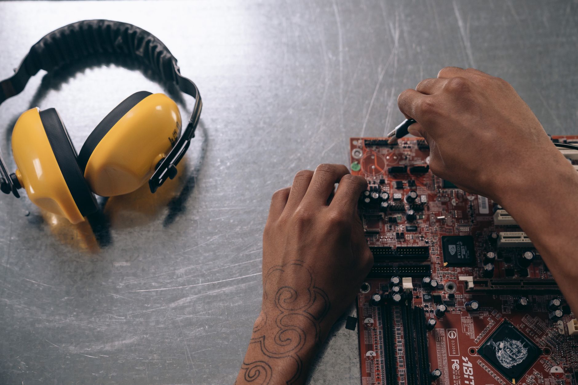 Hiểu Thông Số Loa: Công Suất, Trở Kháng, Độ Nhạy - Hands repairing a motherboard on a metallic surface with yellow earmuffs beside it.