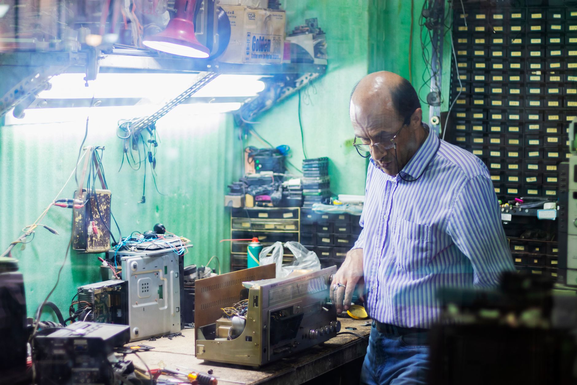 Hiểu Thông Số Loa: Công Suất, Trở Kháng, Độ Nhạy - Man focusing on repairing electronic equipment in cluttered workshop with various tools.